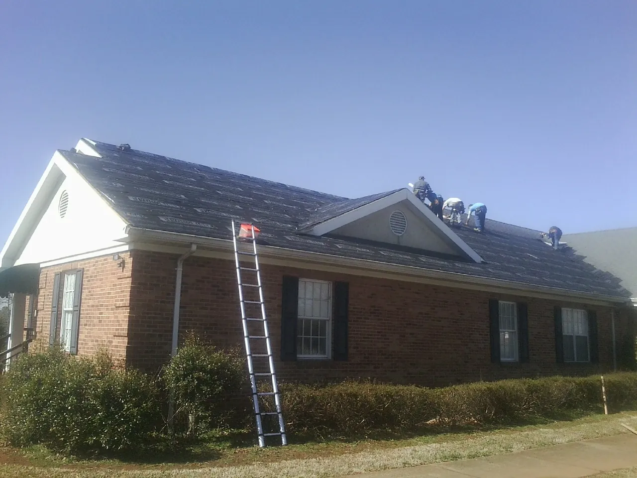 Crew tearing shingles off a brick ranch roof with a ladder set — Metal Roof Repair work by our crew in Blandford
