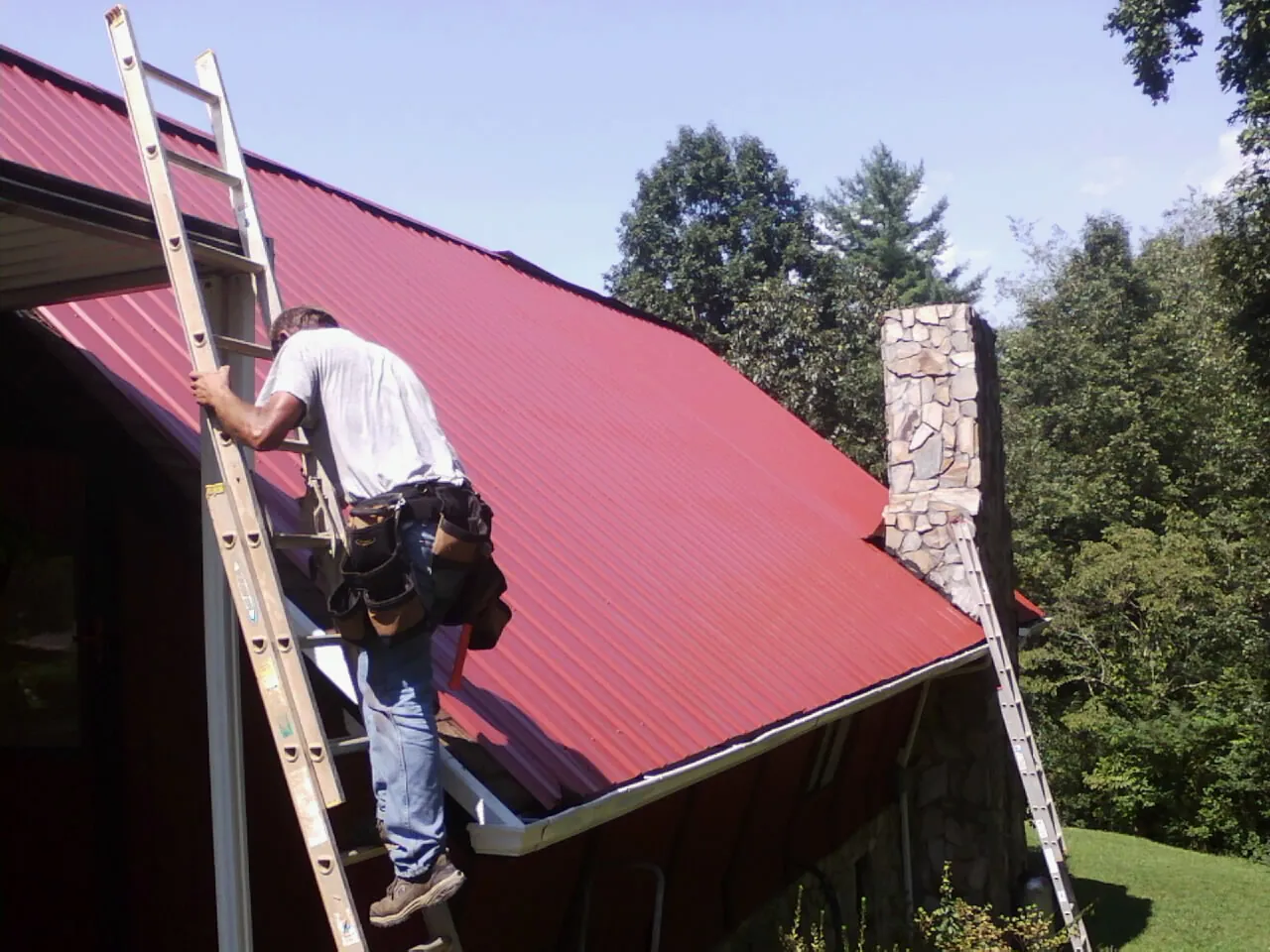 Roofer on a ladder beside a red metal roof and stone chimney — Soffit & Fascia Repair by Roofing Brothers in Blandford